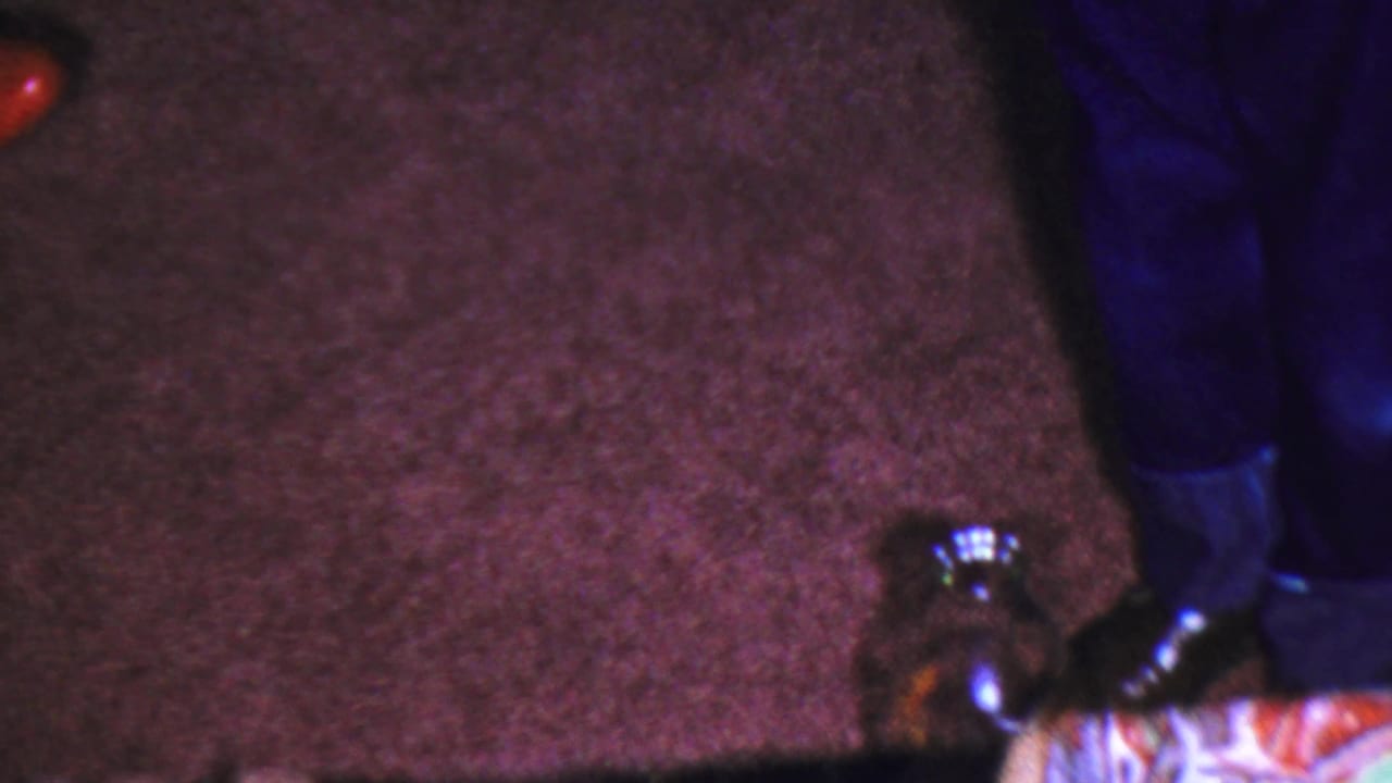 1959 Paramus home-movie frame focused on brown carpeting, lower legs in blue pants, dark shoes, and a glass bottle near the floor.