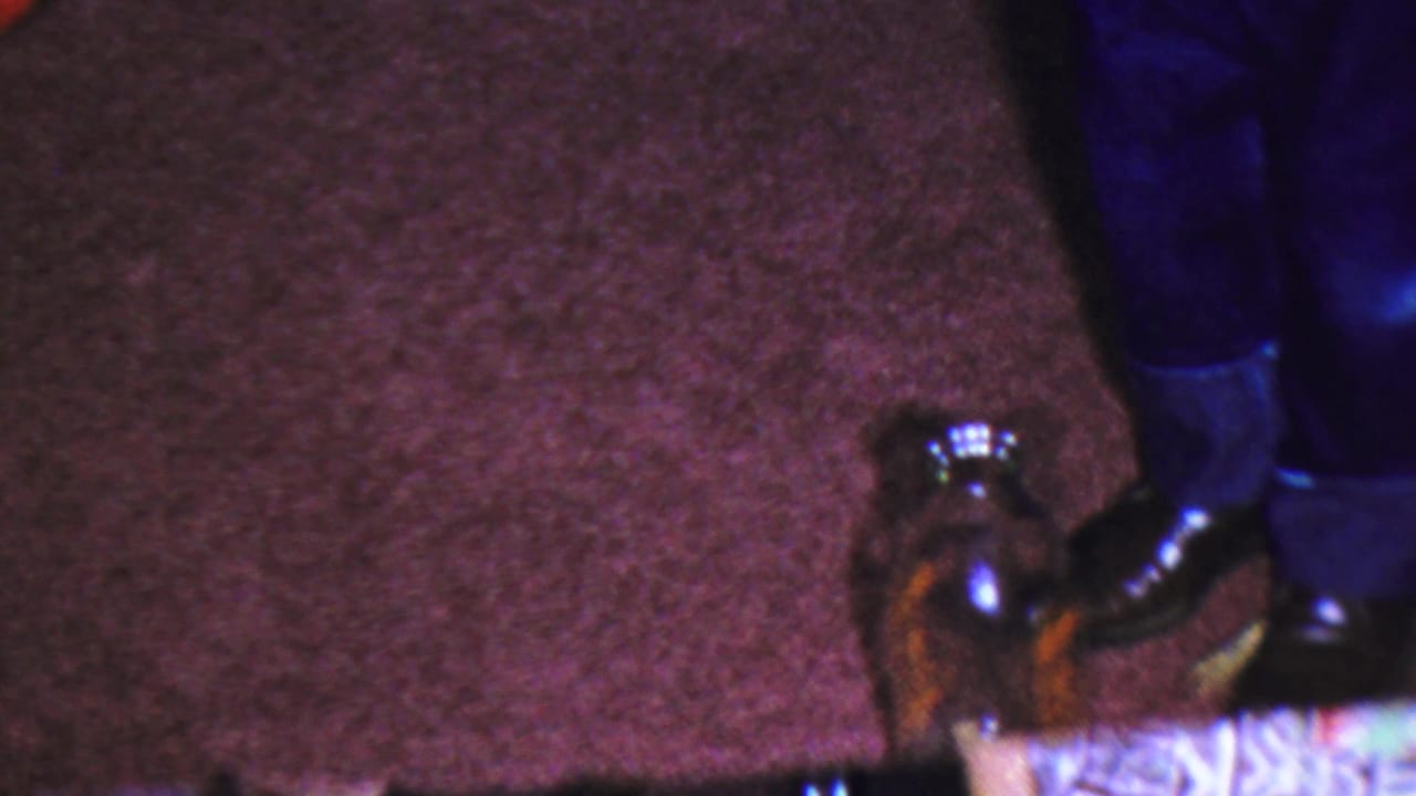 Paramus 1959 8mm frame showing a person's blue pants, polished shoes, brown carpet, and a glass bottle at the lower right of the indoor scene.