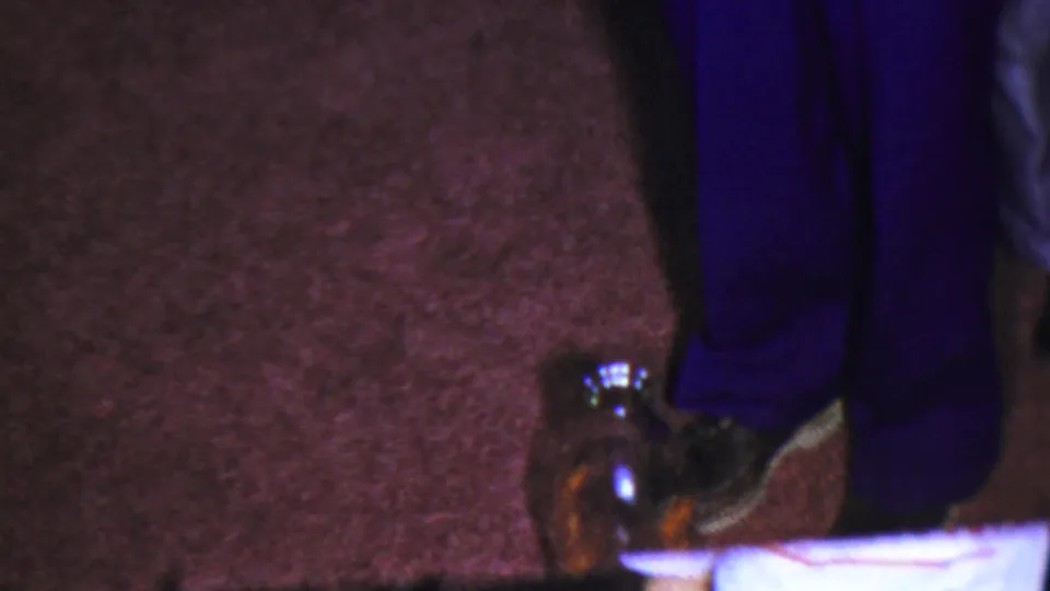 The 8mm frame remains close on the carpeted floor, with the bottle beside black footwear and dark blue trousers.