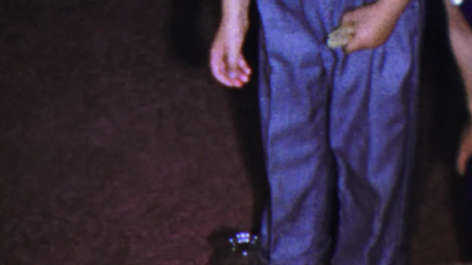 1959 Paramus home-movie frame with cropped adults, polished shoes, maroon carpet, and a dark glass jug on the floor.