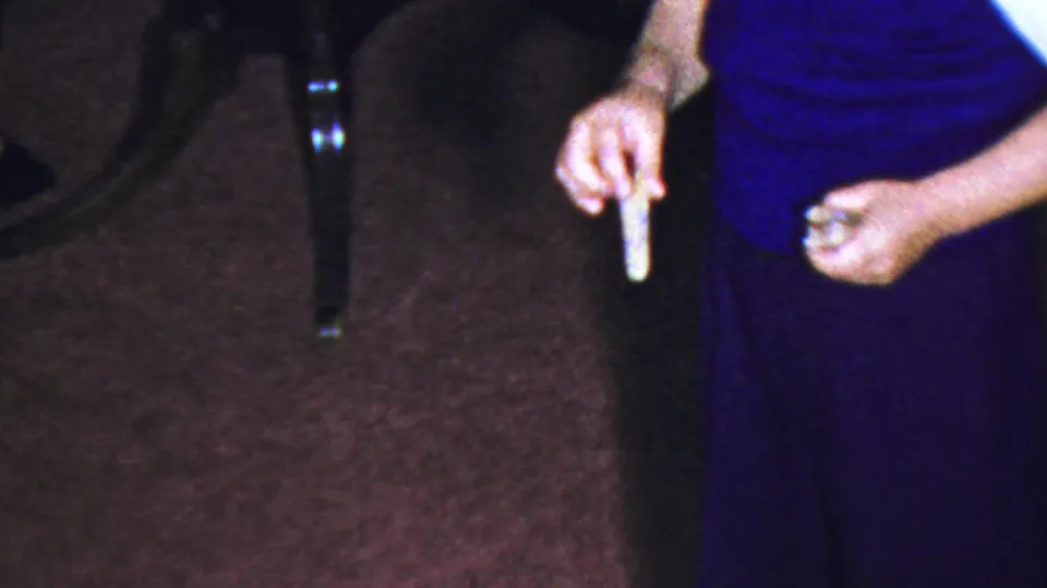 1959 Paramus home-movie frame with an adult in a blue dress holding a cork-like object beside a glass jug on the carpet.