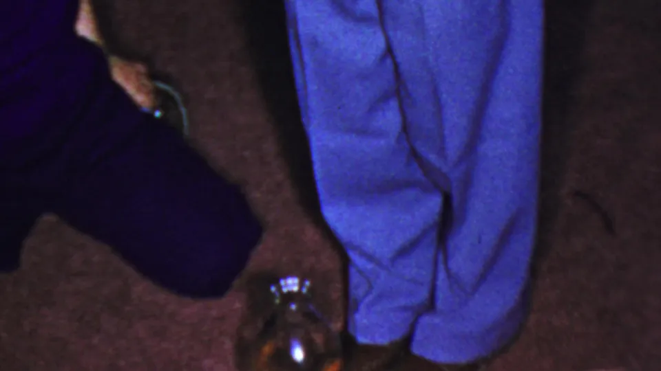 A person in dark clothing bends partly into frame while reaching near the bottle beside the man in blue trousers.