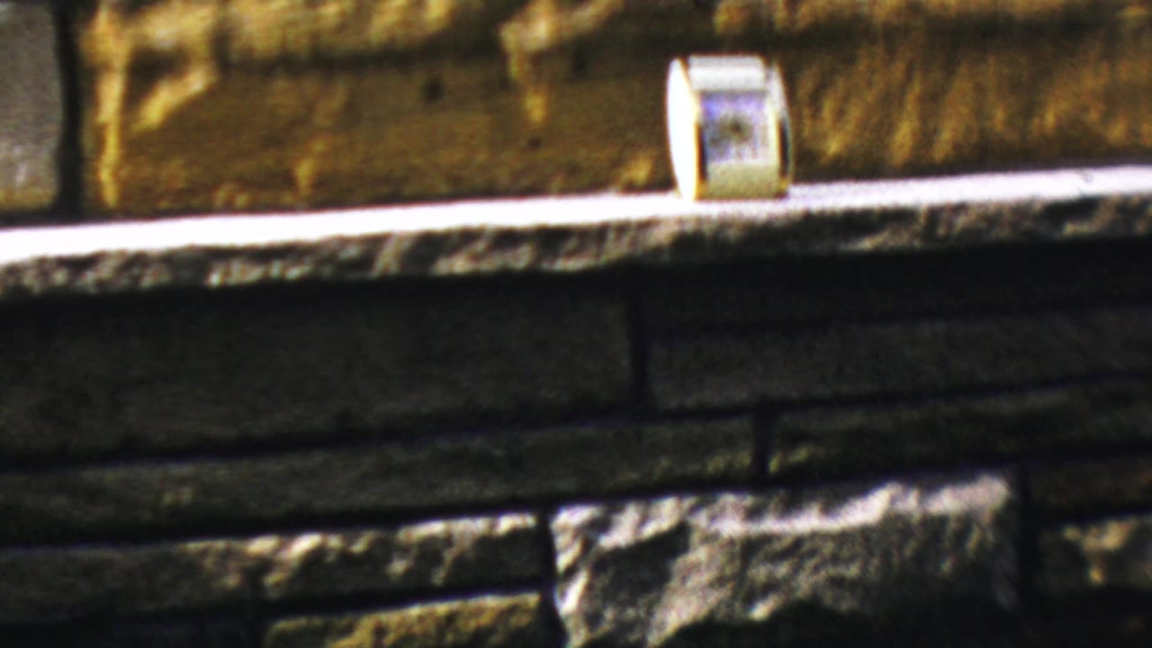 1959 Paramus home-movie frame showing a pale fireplace ledge, stone surround, and a small rectangular item near the hearth.