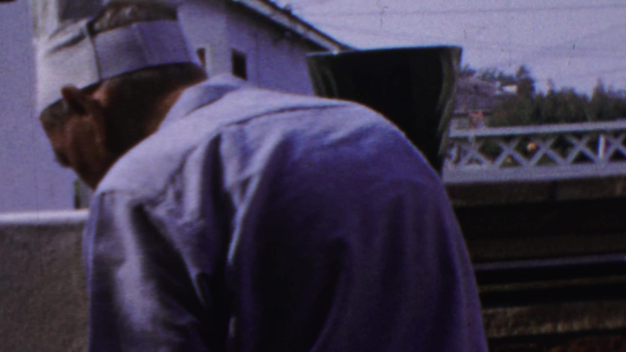 The man turns partly in profile on the Paramus patio, wearing a white chef hat and light shirt near the outdoor cooking area.