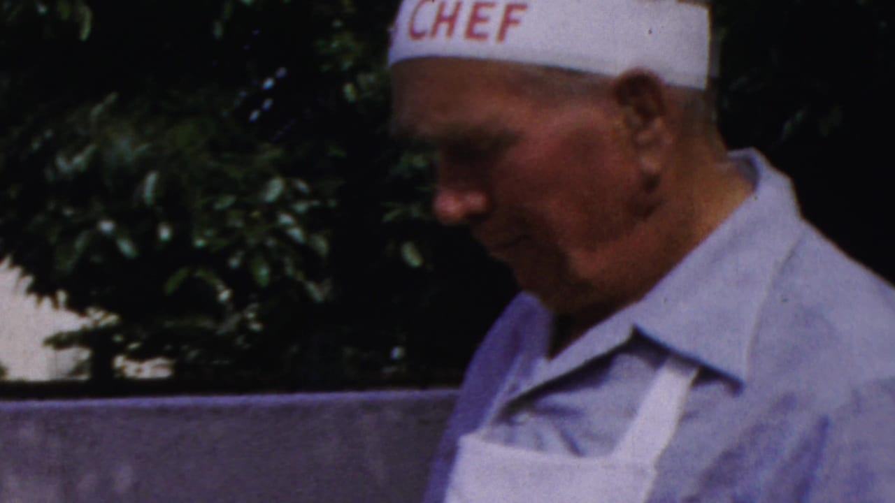 Man in a white chef hat and apron stands beside an outdoor grill.