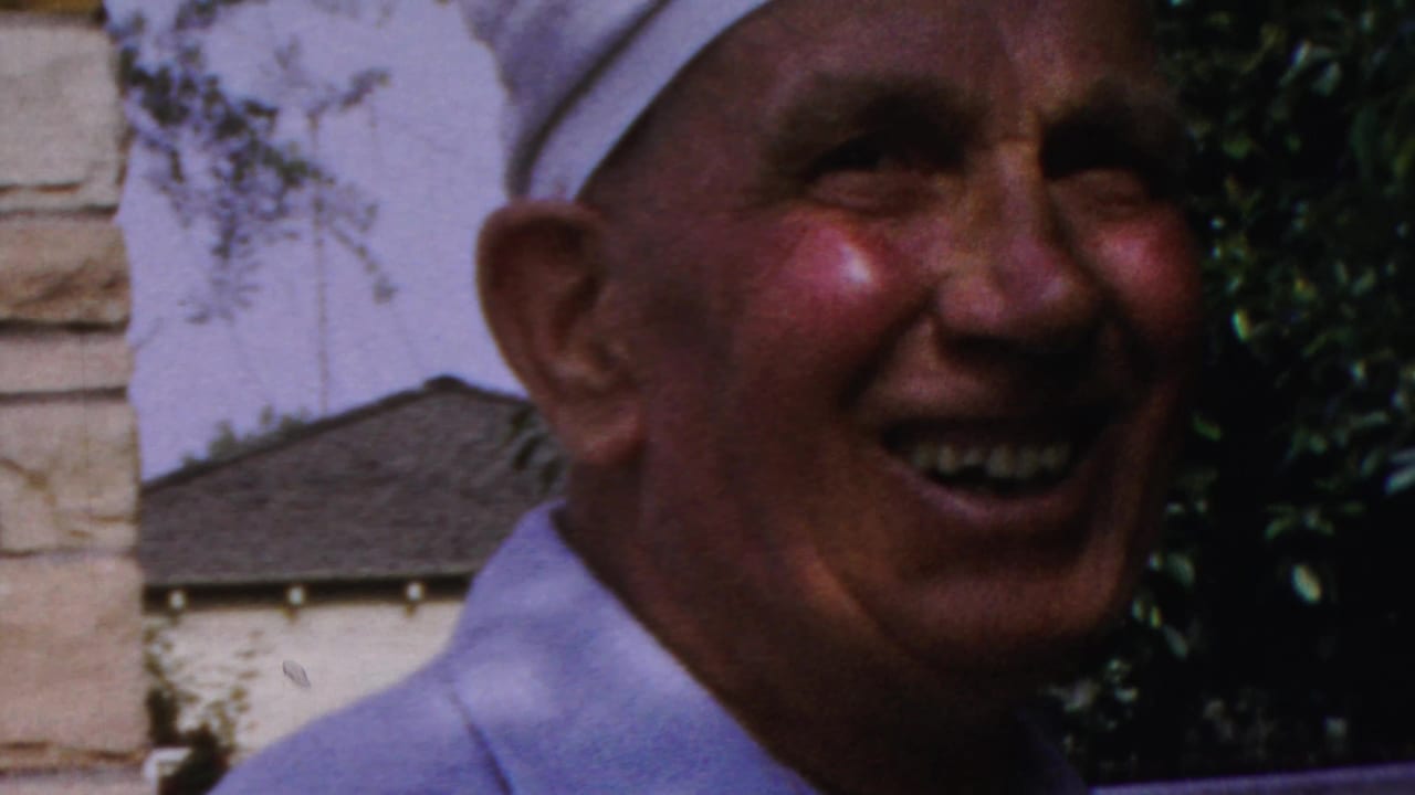 1959 Paramus home-movie frame showing the man smiling broadly in front of foliage and a stone wall.