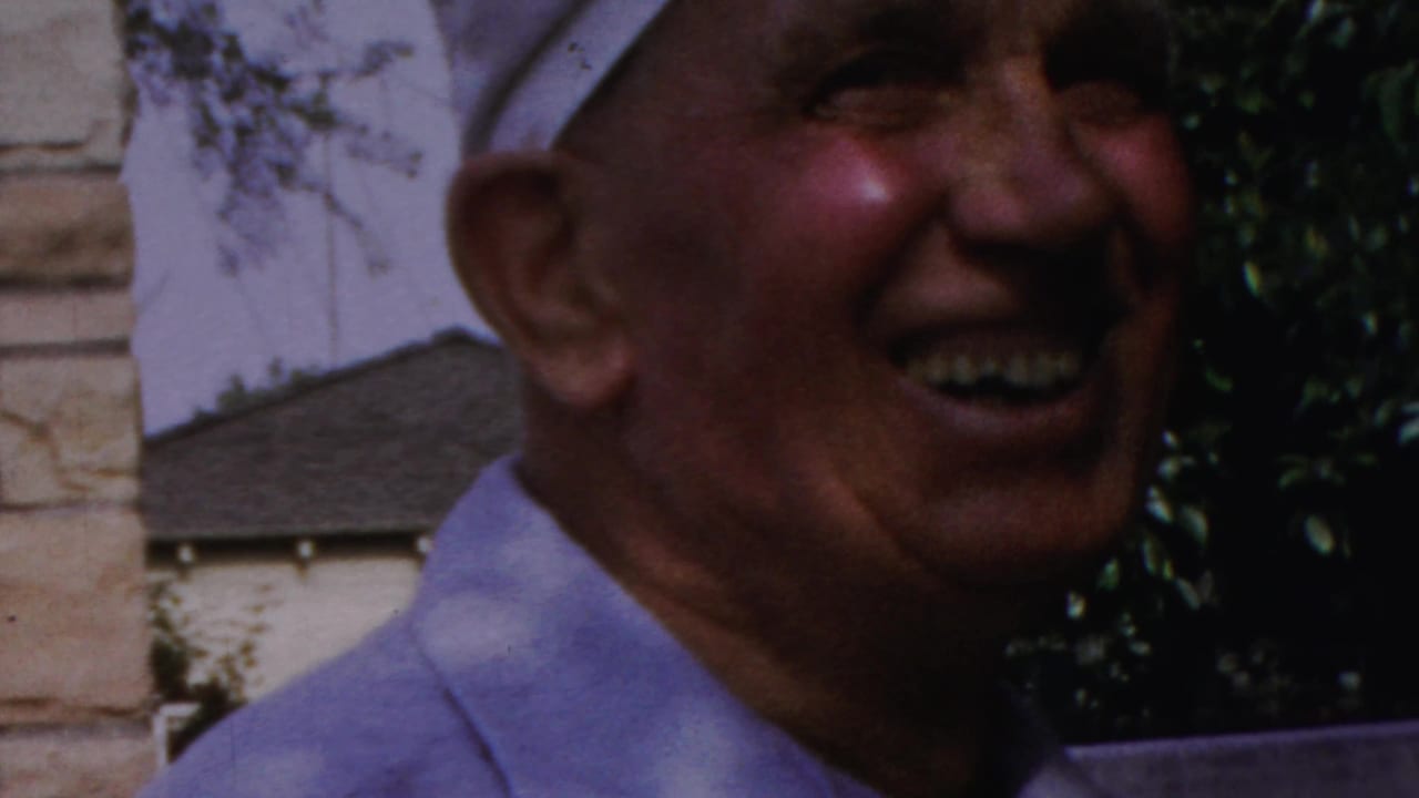 1959 Paramus frame showing the man in a white cap laughing or smiling widely near shrubs and stonework.