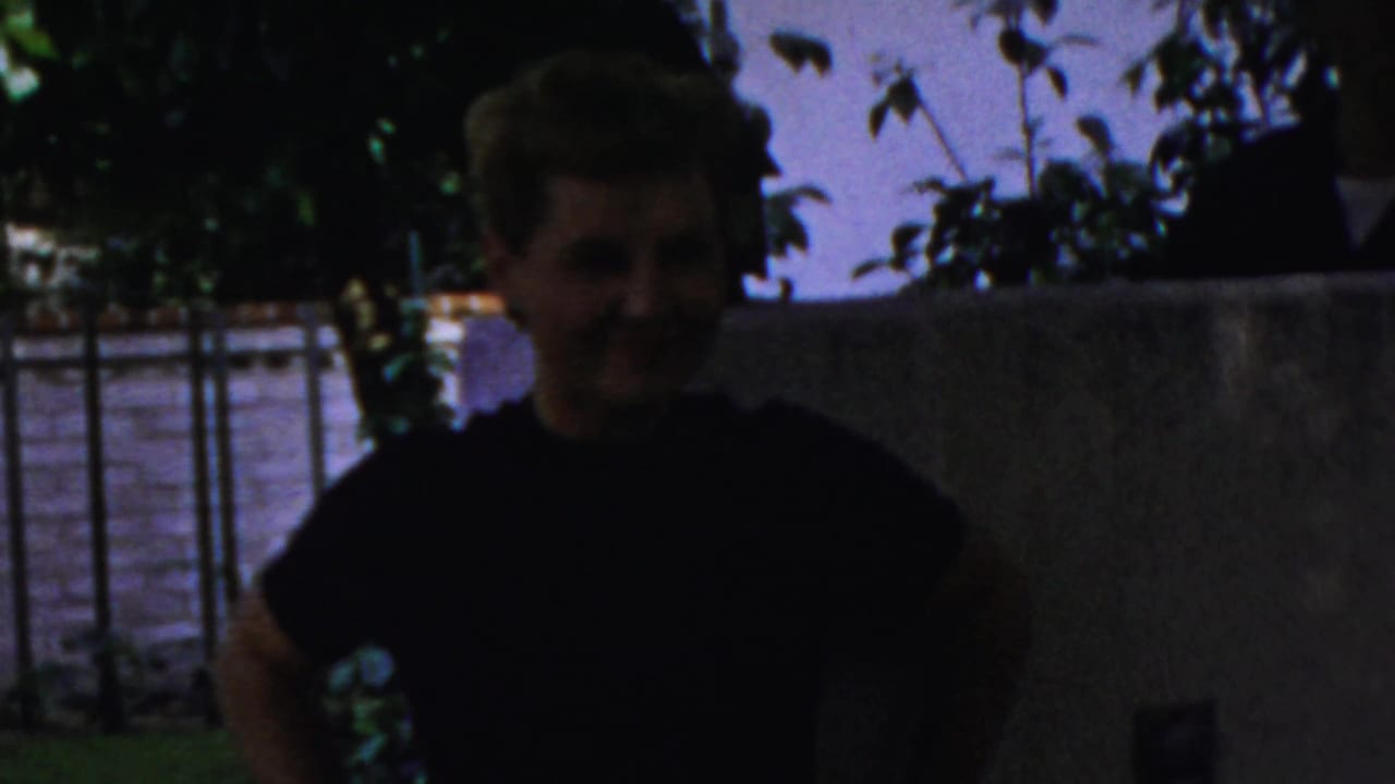 In 1959 Paramus home-movie footage, a young woman with short hair stands near a low wall in a shaded backyard.