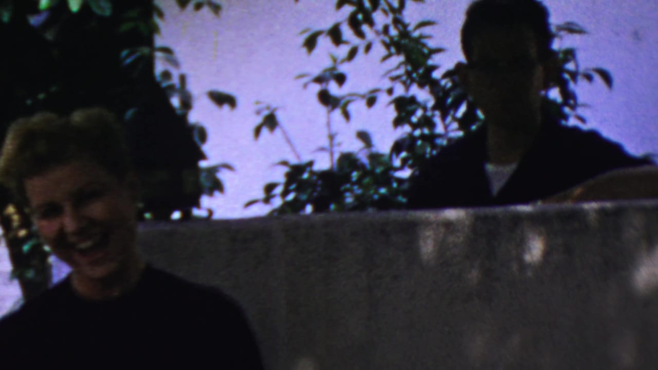 A young woman laughs in the foreground as an older man stands partly silhouetted behind a backyard wall in Paramus, 1959.