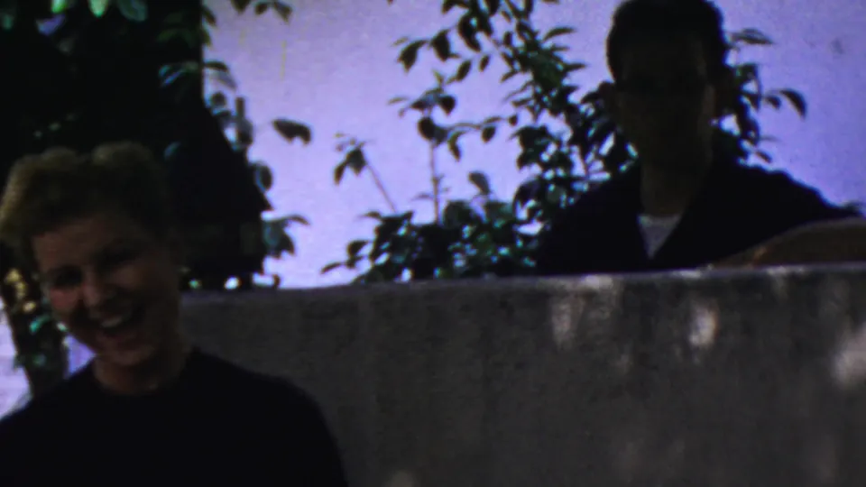 A young woman laughs in the foreground as an older man stands partly silhouetted behind a backyard wall in Paramus, 1959.