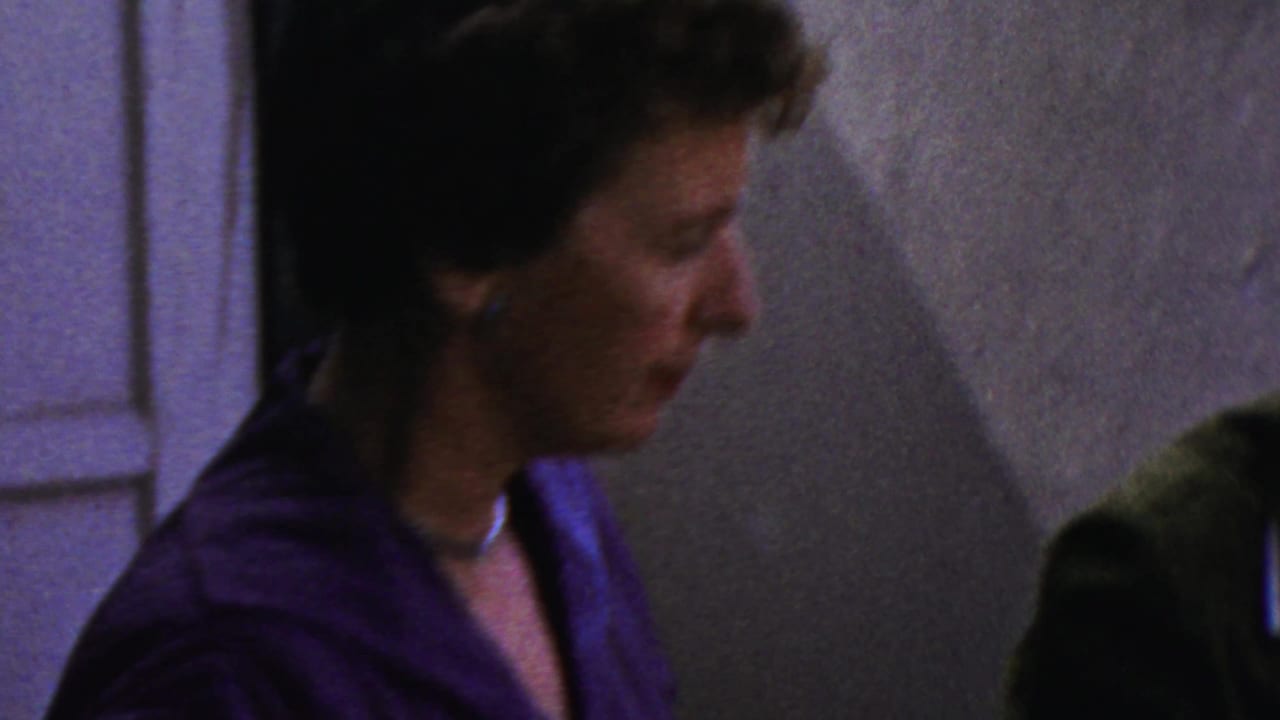 Woman in a blue-purple dress seen in profile inside a home near a serving table.
