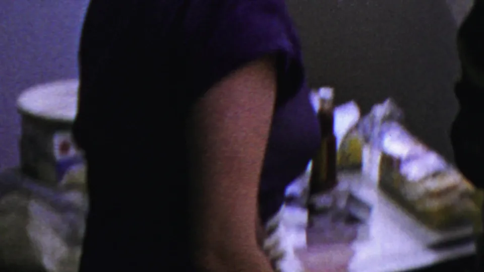 The frame shows a cropped adult figure in a blue-purple dress near a serving table, with another person partly visible.