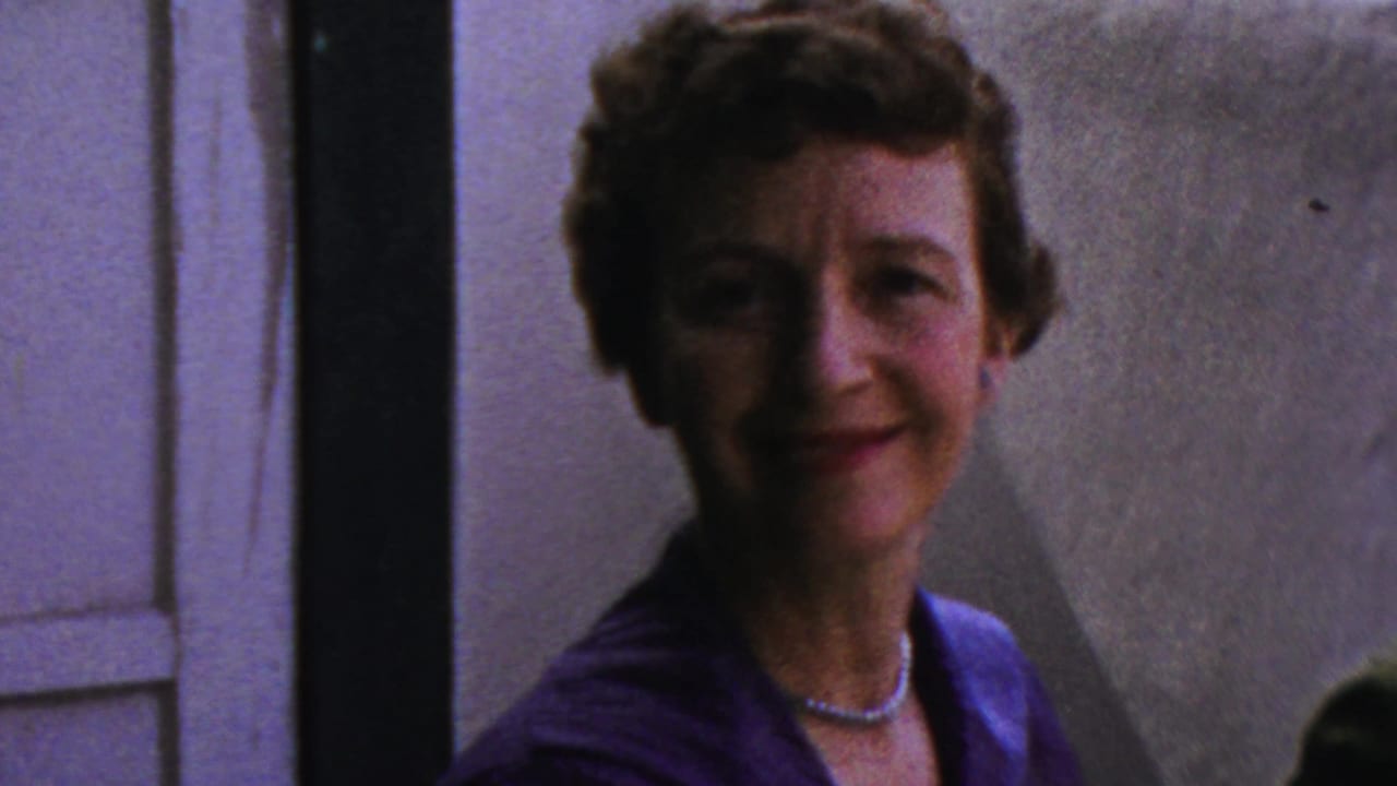 A woman in a blue-purple dress and pearl necklace smiles toward the camera in 1959 Paramus home-movie footage.