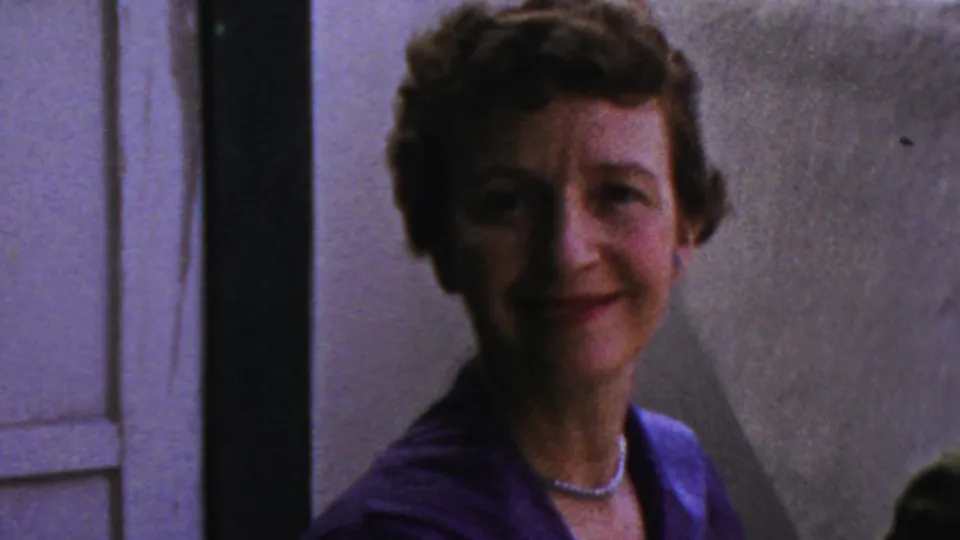 A woman in a blue-purple dress and pearl necklace smiles toward the camera in 1959 Paramus home-movie footage.