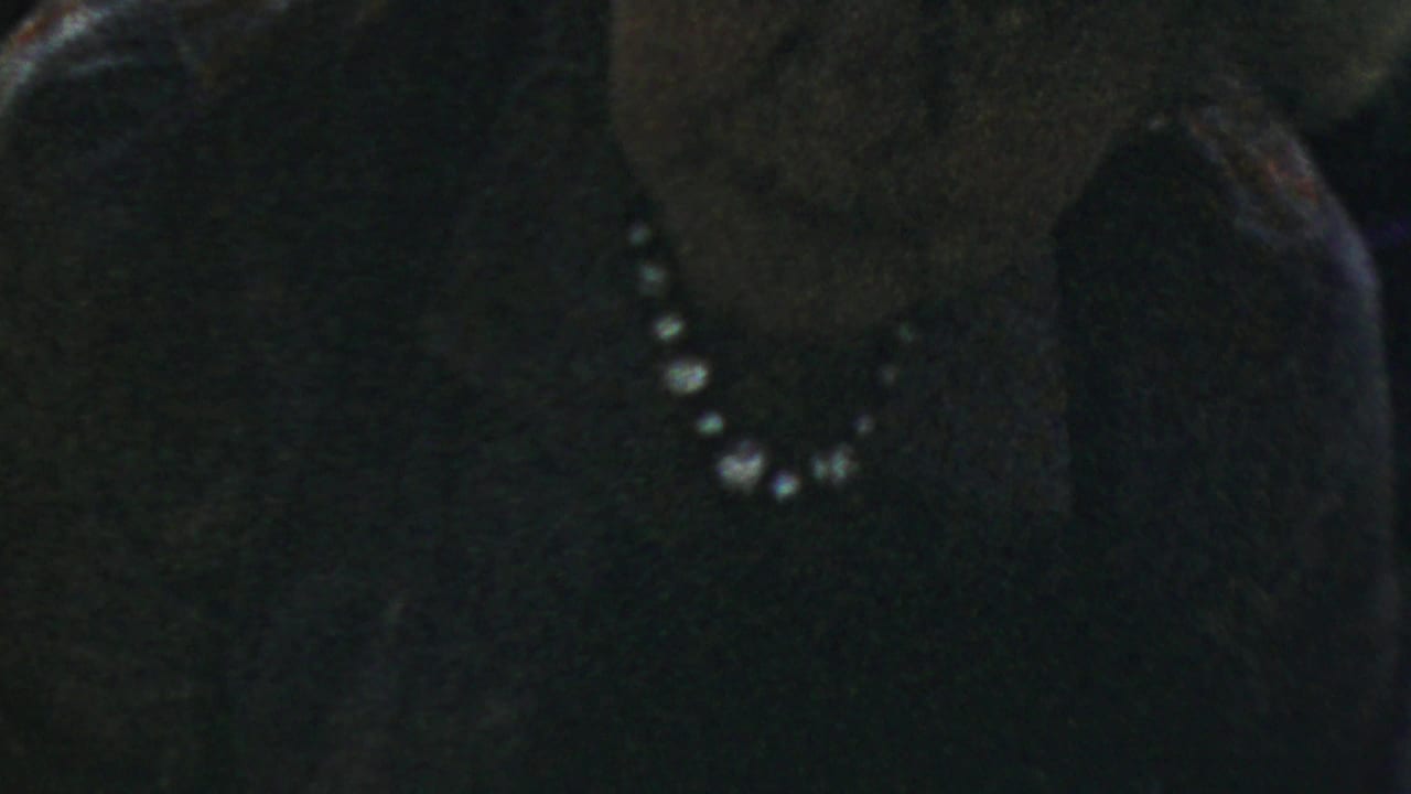 A close 8mm frame shows the woman with glasses and pearls, her face softly visible against a dark setting.