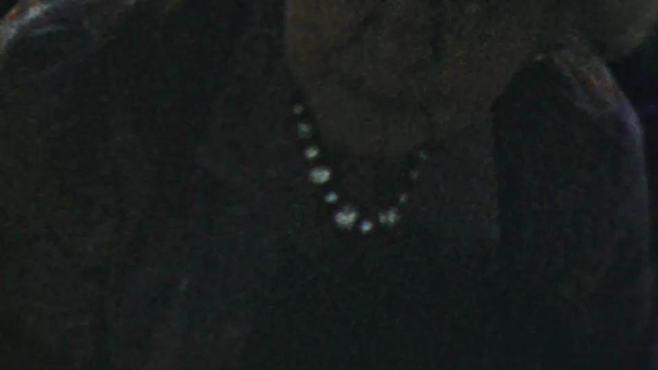 A close 8mm frame shows the woman with glasses and pearls, her face softly visible against a dark setting.