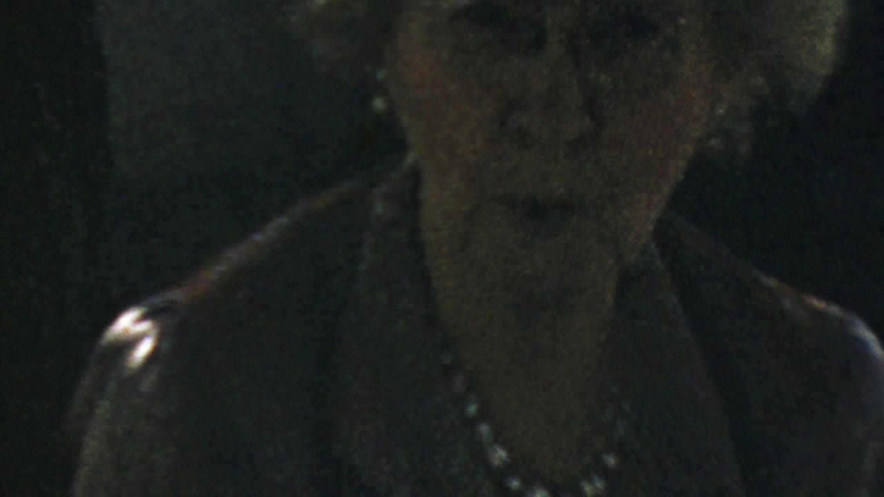 1959 home-movie frame of the woman looking nearer to the lens, with glasses, pearls, and dark clothing visible.