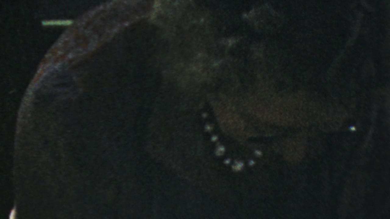 Woman in a dark dress and pearl necklace seated in dim interior light.