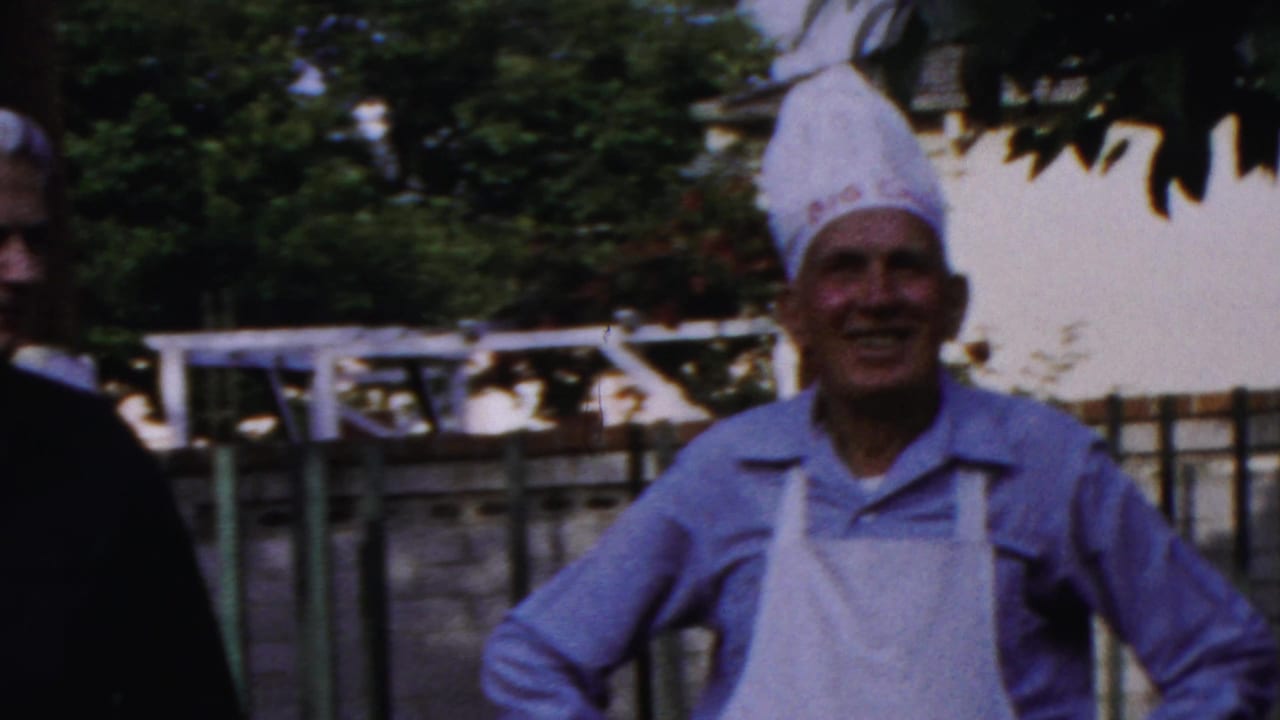 1959 Paramus home-movie frame showing adults at an outdoor barbecue, including two men in chef hats and aprons near a fence.