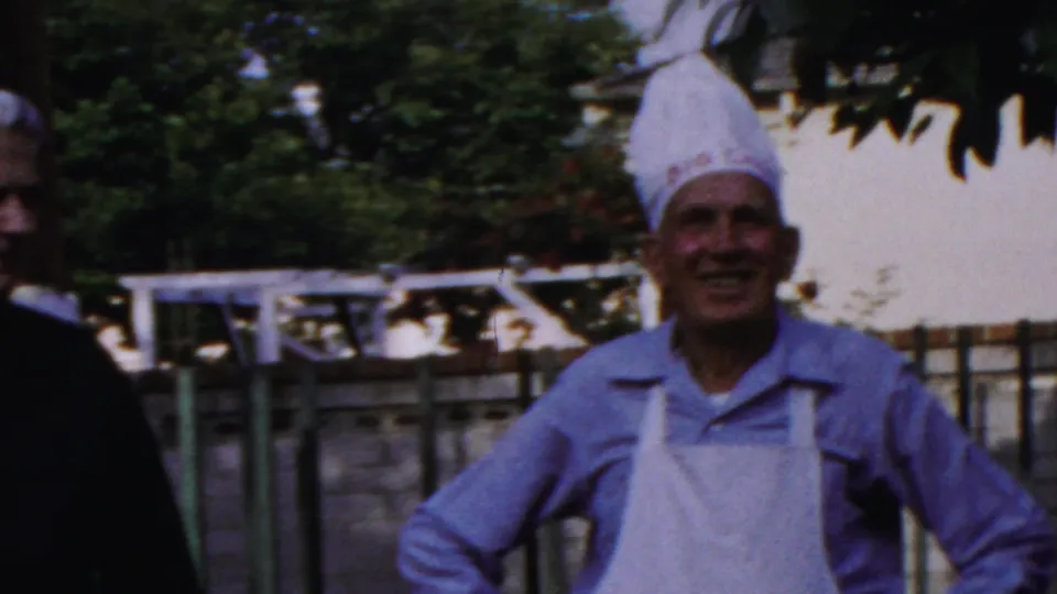 1959 Paramus home-movie frame showing adults at an outdoor barbecue, including two men in chef hats and aprons near a fence.