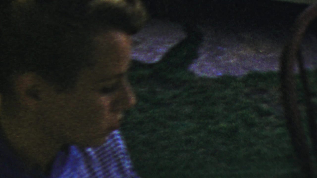 1959 Paramus home-movie frame showing a young person in a checkered shirt beside chairs and grass near a meal area.