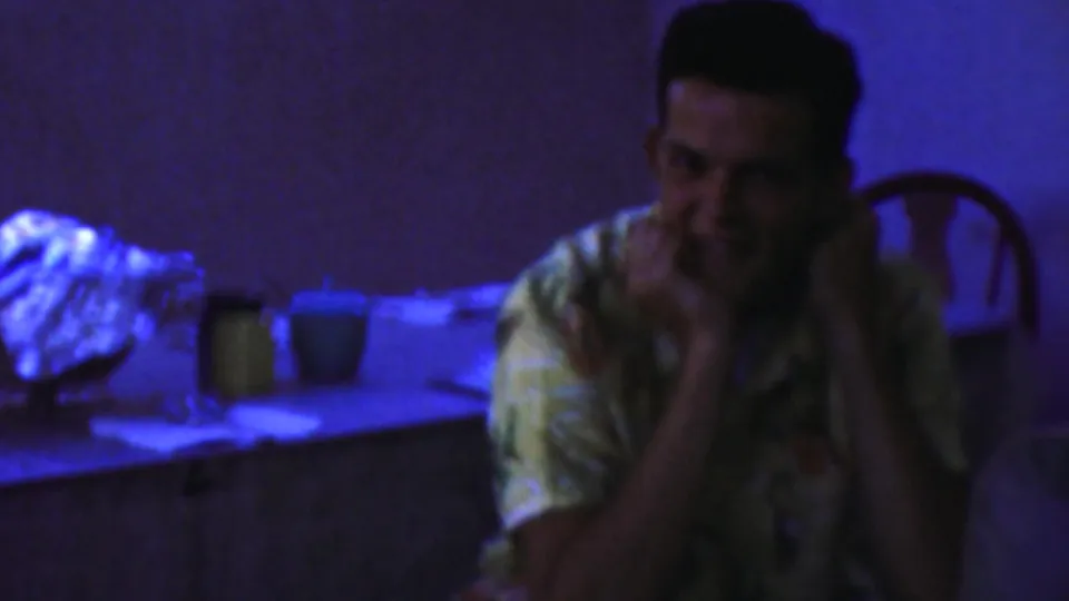 A seated man in a patterned shirt smiles with his hands near his face in the 1959 Paramus indoor gathering footage.