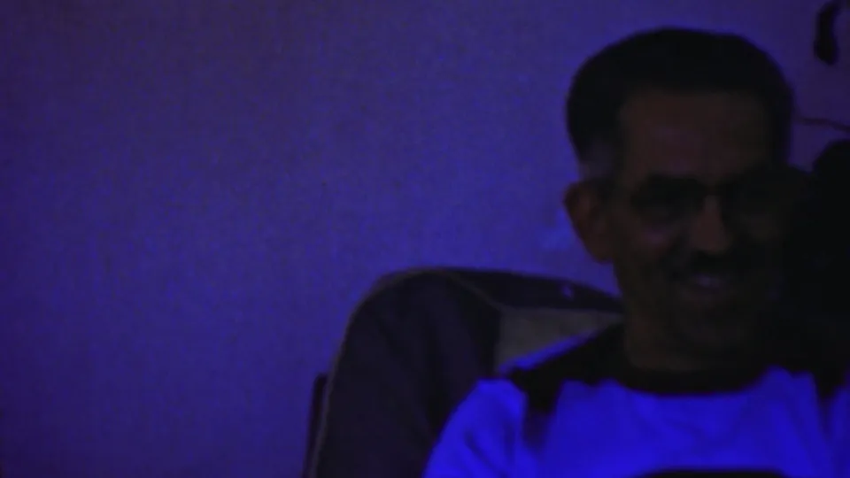 A man wearing glasses smiles toward the camera in a blue-purple private interior, identified by metadata as 1959 Paramus footage.