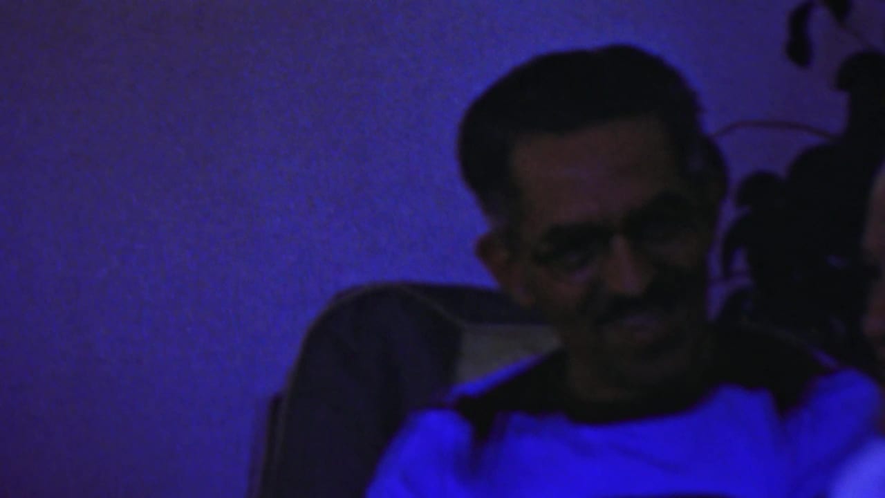 Adult man seated indoors in Paramus home-movie footage, lit by intense blue light in a dark room.