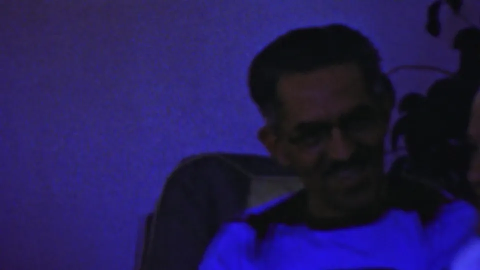 Adult man seated indoors in Paramus home-movie footage, lit by intense blue light in a dark room.