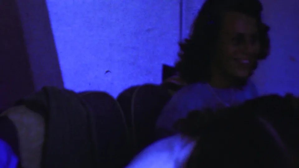 An adult woman smiles in a dim private room while other adults sit nearby in blue-toned 8mm footage.