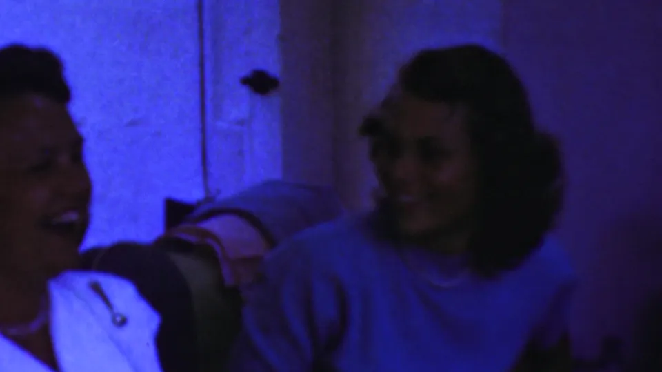 Two adult women appear to laugh together in a blue-lit private interior from Paramus, New Jersey home-movie footage.