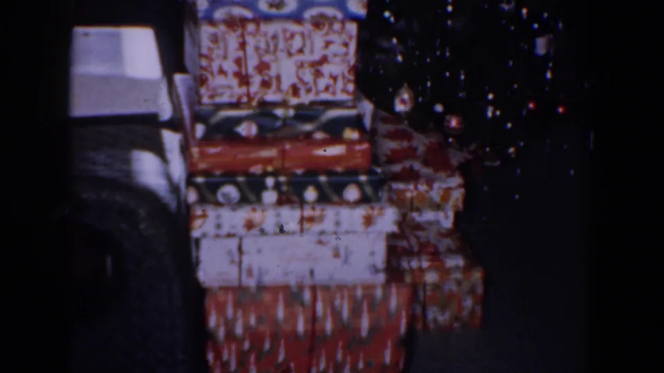 1960 Bel Air, Maryland home-movie frame showing wrapped gifts stacked beside a decorated Christmas tree in a dim room.