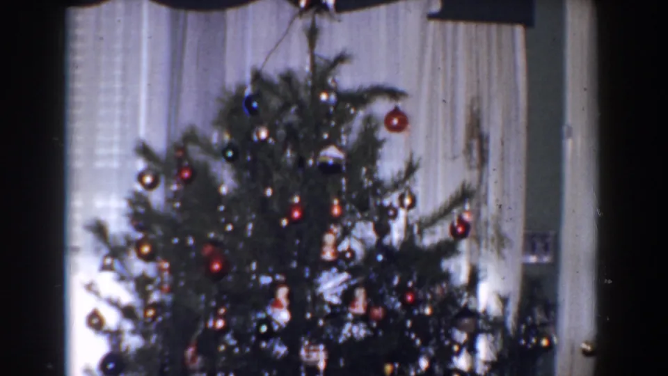 1960 Bel Air, Maryland home-movie frame showing a Christmas tree with red star topper, hanging ornaments, tinsel, curtains, and window blinds.