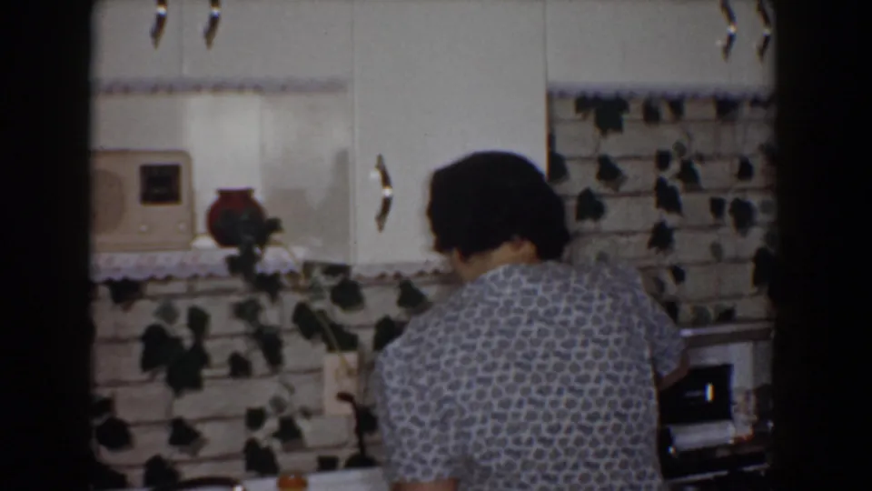 1960 Bel Air, Maryland frame of a woman seen from behind cooking at the stove beneath white cabinets and ivy-decorated backsplash.
