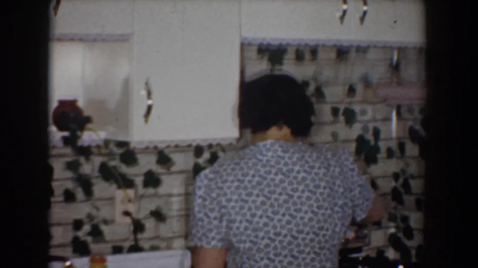 Home-movie frame from the Bel Air kitchen showing the woman leaning at the stove, with white cabinets, backsplash, and counter details visible.