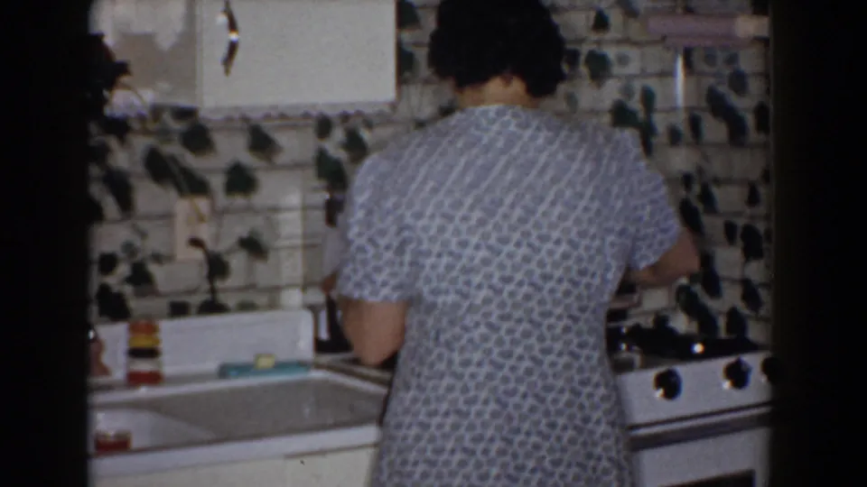 1960 Bel Air, Maryland frame showing the woman at the stove beside a sink, with white cabinets and ivy-decorated backsplash in view.