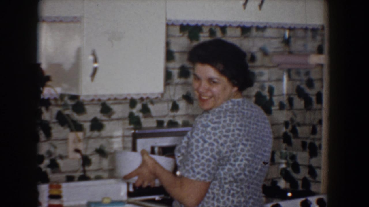 In a 1960 Bel Air, Maryland home-movie frame, a woman smiles toward the camera while holding a white bowl near a kitchen counter.