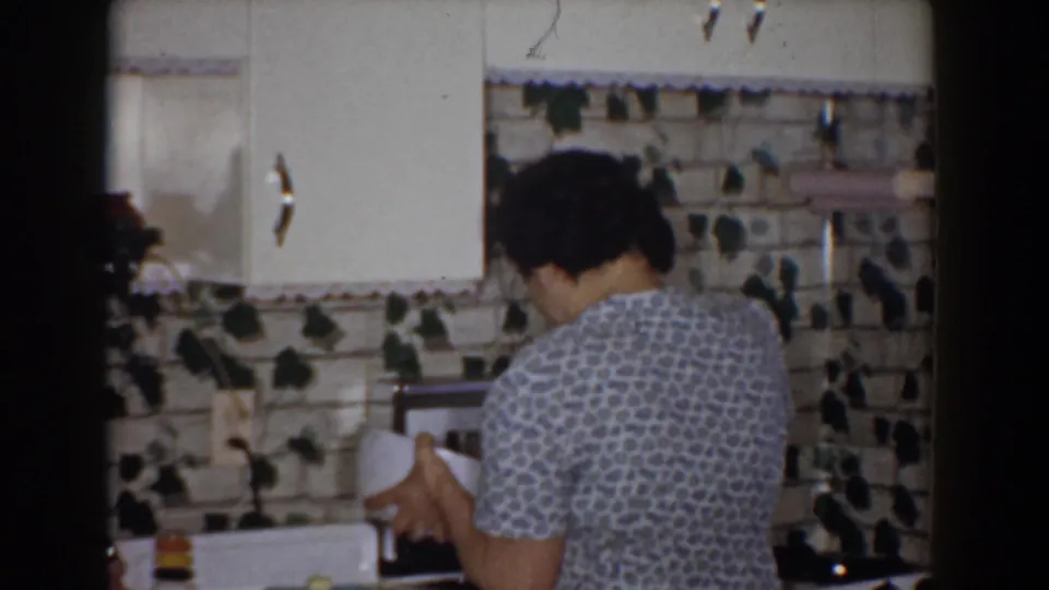The woman faces away from the camera while holding a white bowl at the counter in the 1960 Bel Air kitchen scene.