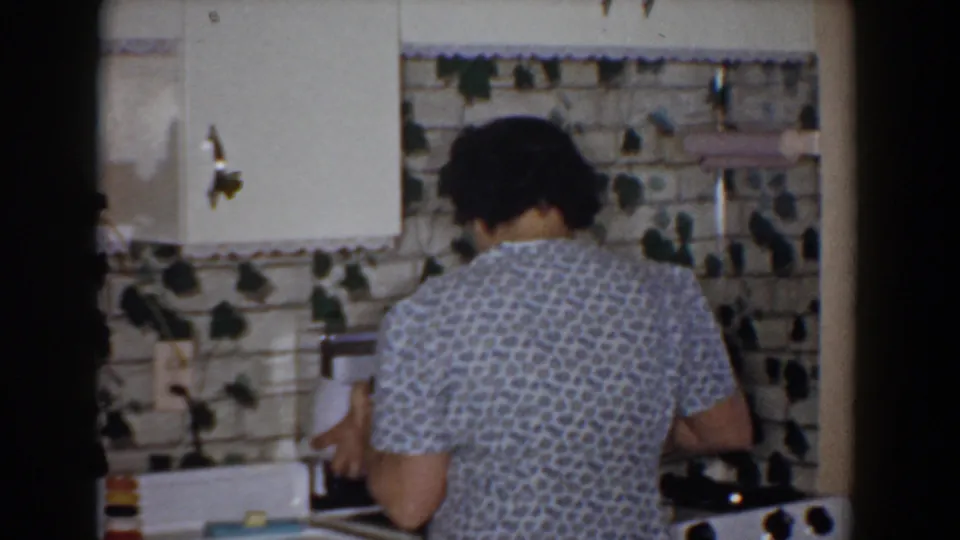 A 1960 home-movie frame shows the woman from behind as she works at the kitchen counter with a white bowl in hand.
