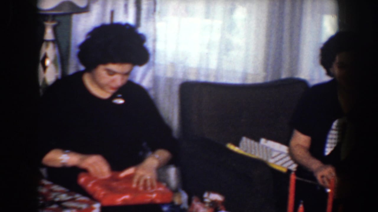 Woman in black seated indoors, unwrapping a red gift beside curtains and a chair.