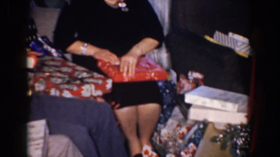 A woman seated in a private living room holds a red Christmas gift, with packages and furniture around her.