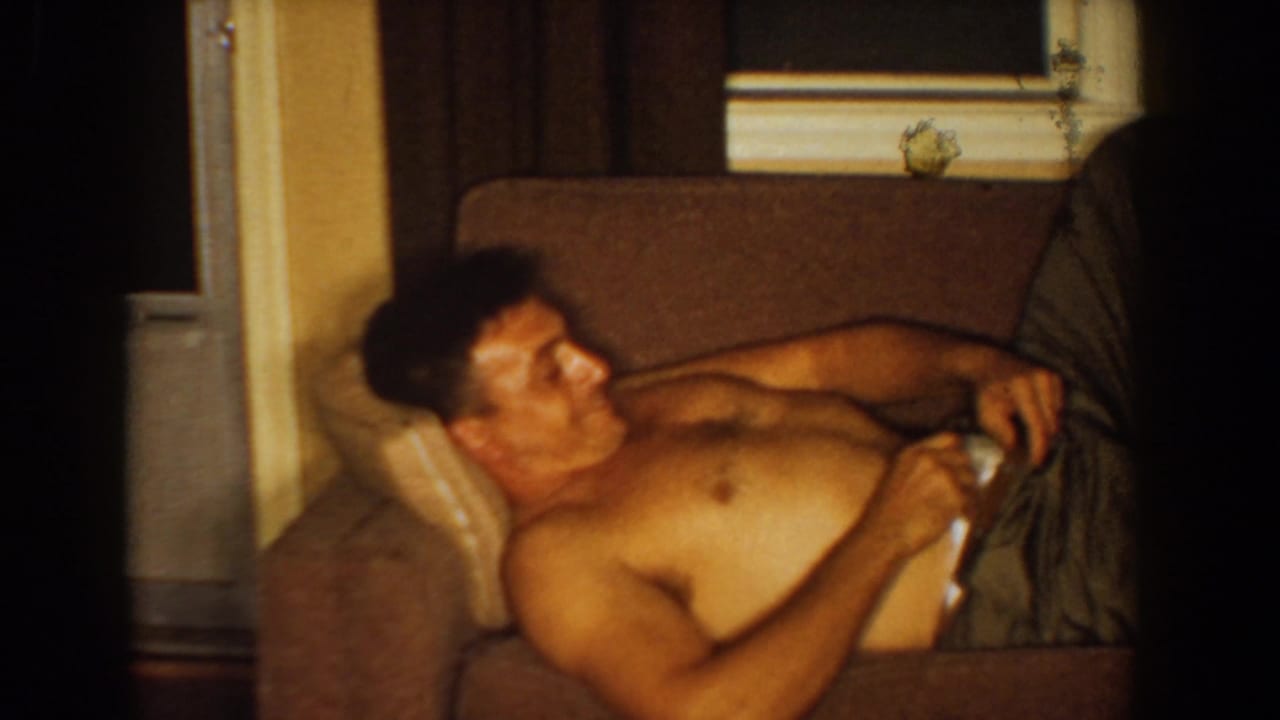 Shirtless man reclining on a brown couch in a dim living room.
