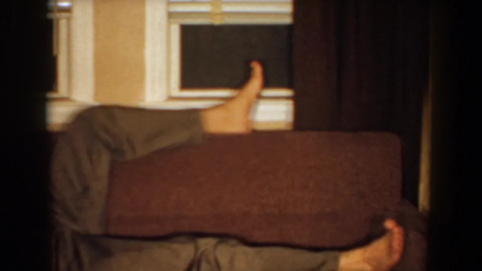 The camera catches the couch back with the reclining man's bare feet visible near the curtained window.