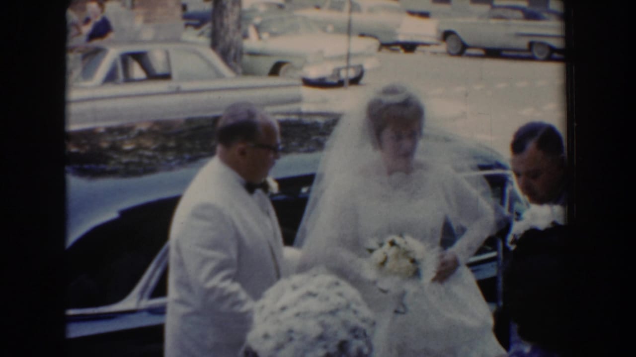 Bride holds a bouquet beside a car while a man in a white jacket assists her.