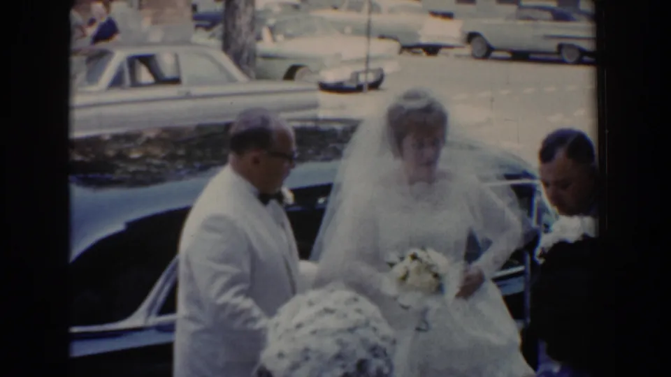 The bride holds her bouquet beside the curbside car, assisted by a man in formalwear in the 1960 wedding scene.