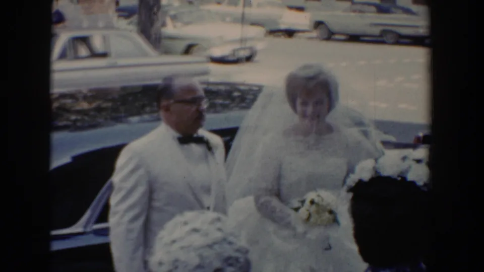 The bride and a man in a white tuxedo jacket stand at the curb beside a dark car during the 1960 wedding arrival.