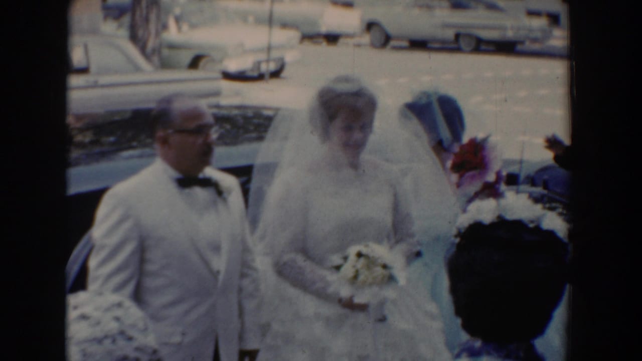 Bride and father stand near a bridesmaid holding flowers beside parked cars.