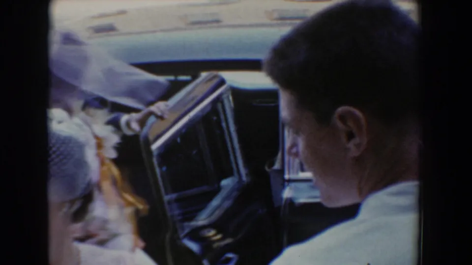 An open car door and crowded wedding guests frame the bride's movement away from the vehicle during the 1960 home-movie scene.