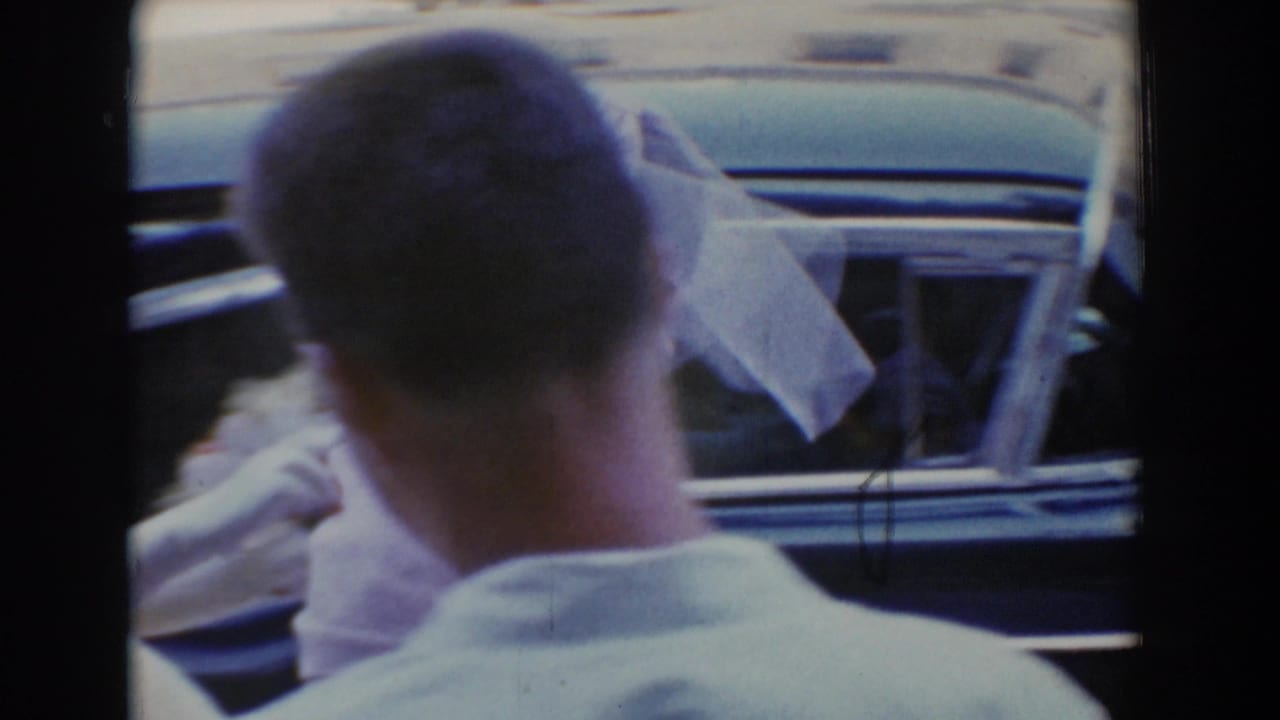 Man seen from behind near a car as a veiled bride stands outside.