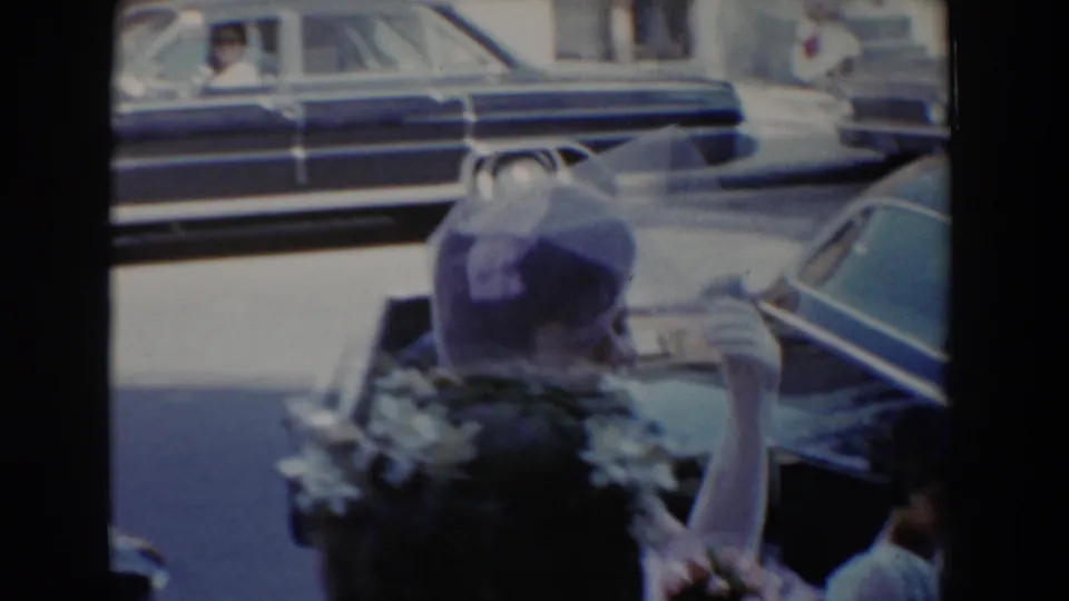 Bel Air, Maryland 1960 wedding frame showing a woman in a formal hat holding flowers near parked cars.